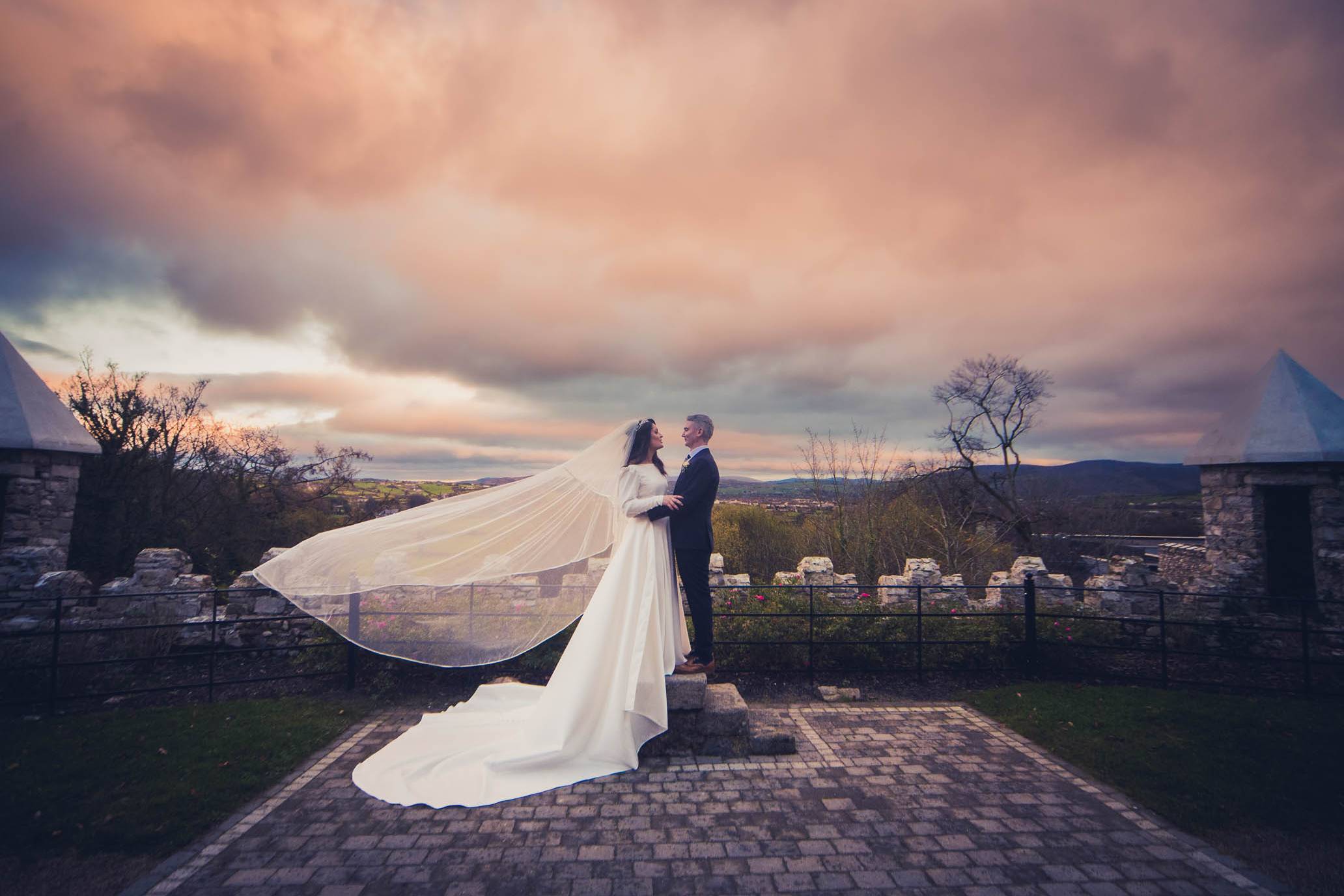 Birde and groom standing outside Killeavy Castle with her veil flowing in the wind