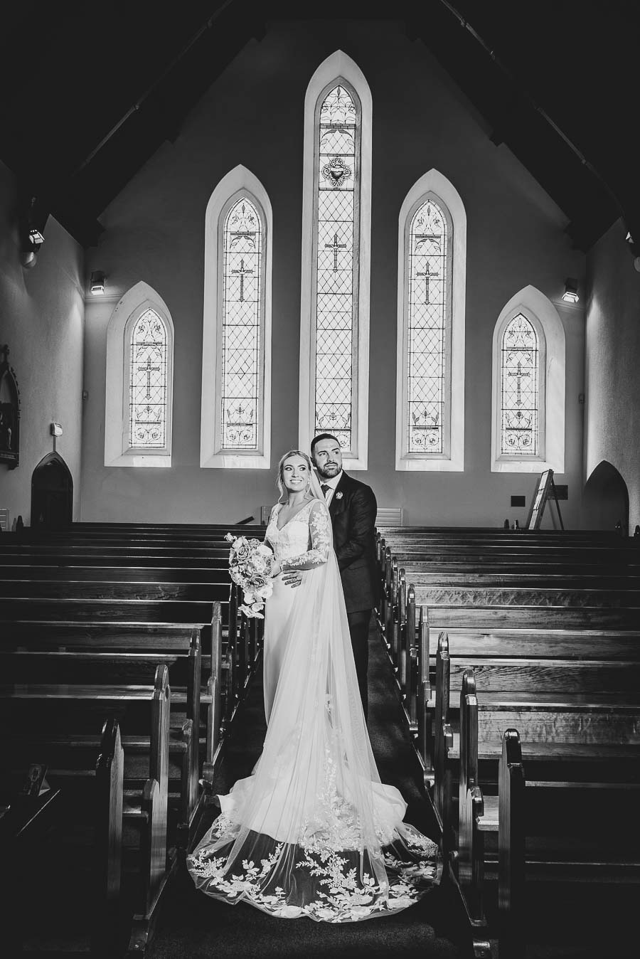 Melissa & Aidan standing in the aisle with beautiful stain glassed windows in background