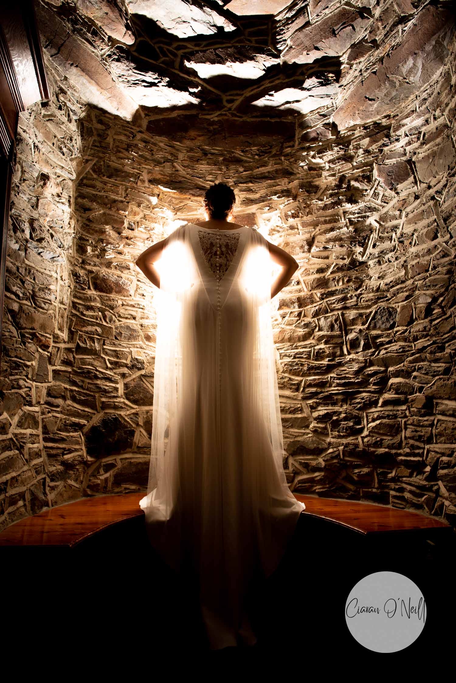 Bride standing in front of old stone wall facing away from the camera with light shining through the dress