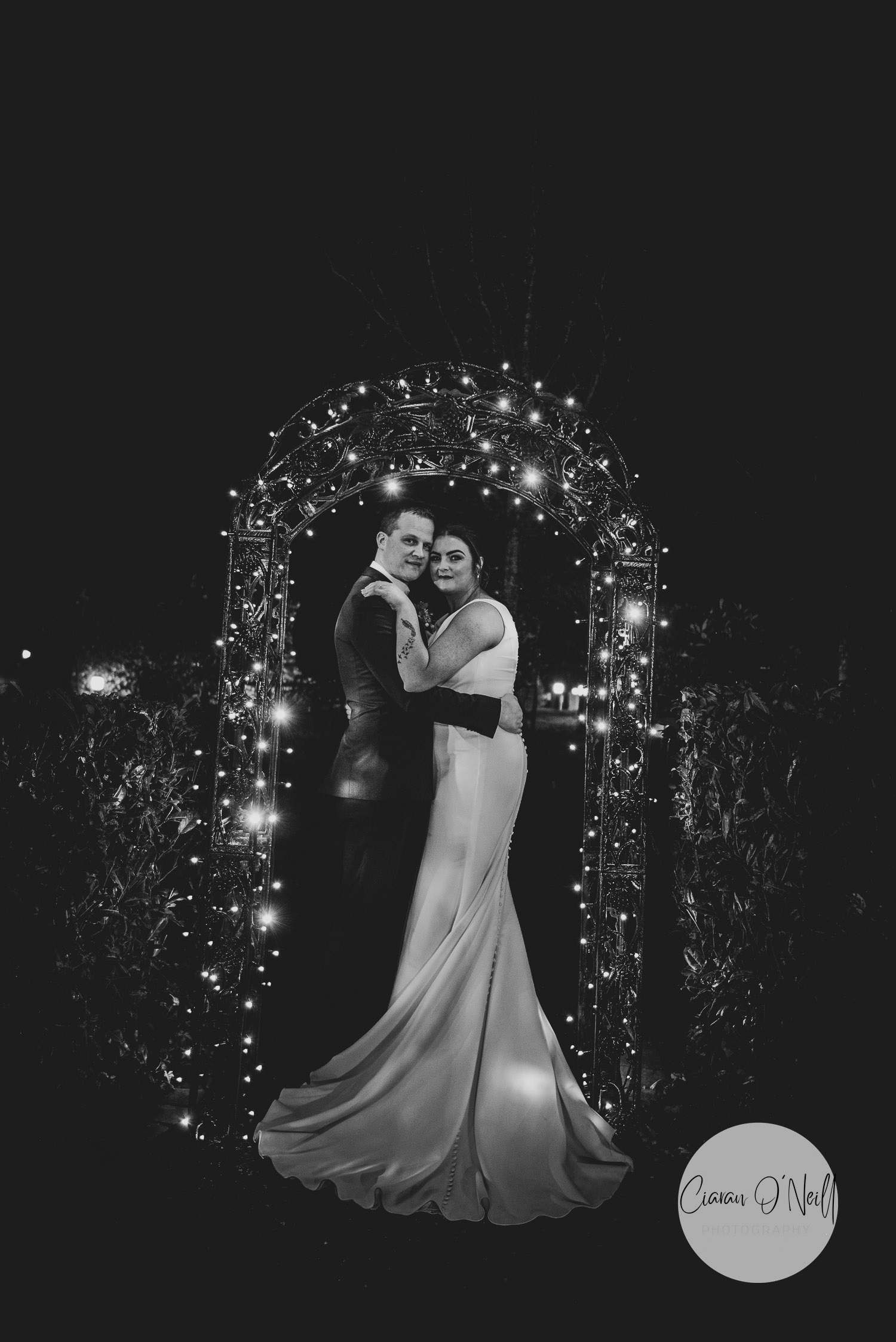 nighttime photo of bride and groom lit with fairy lights on an archway