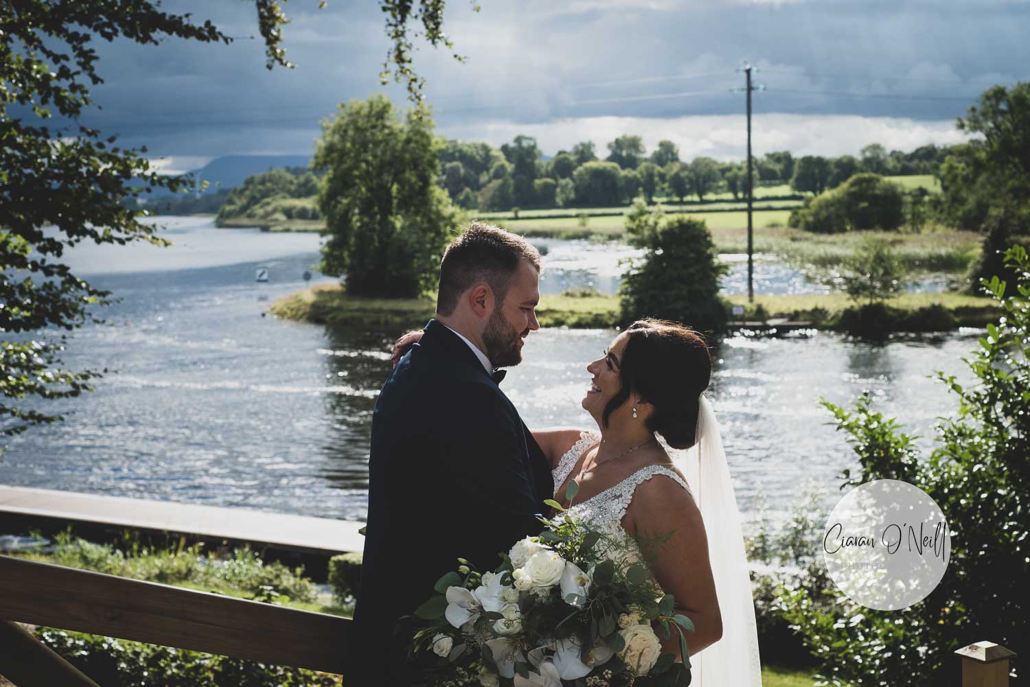Bride and groom embracing in front of a sunlit lake at the Killyhevlin Hotel