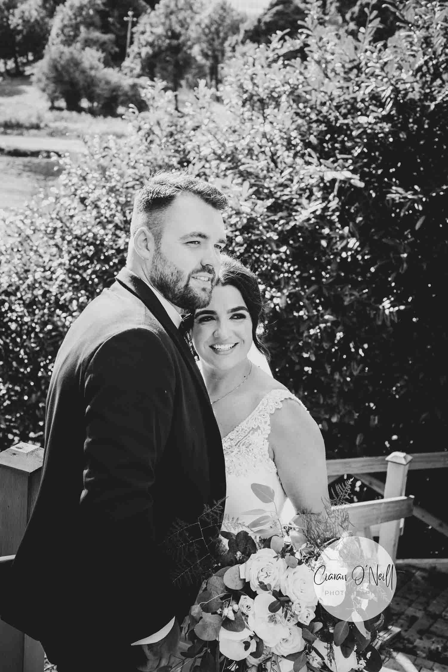 Black and white photo of bride and groom looking off to one side in front of wooden fence and trees