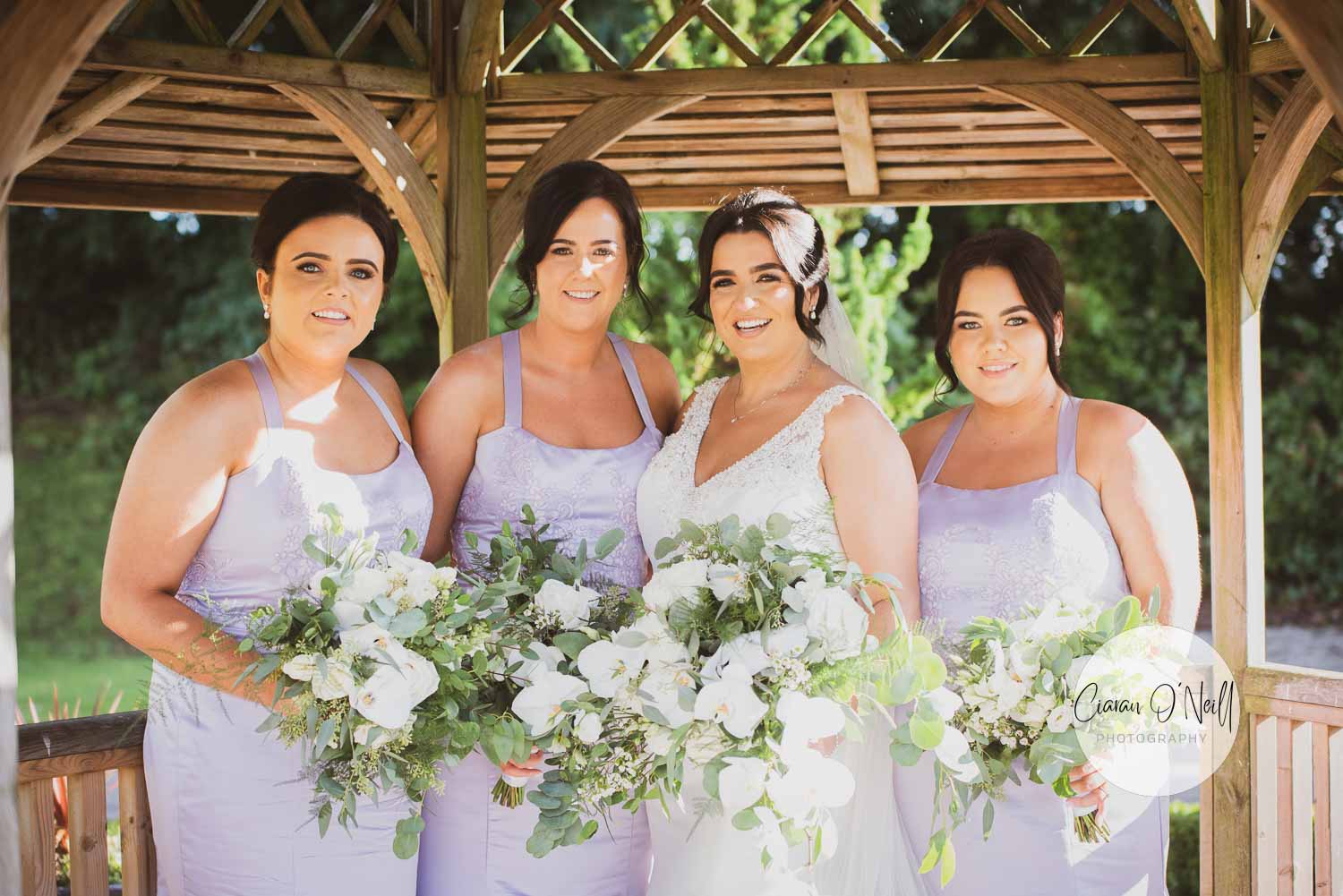 bRide and three bridesmaids wearing lilac dresses under a wooden gazebo