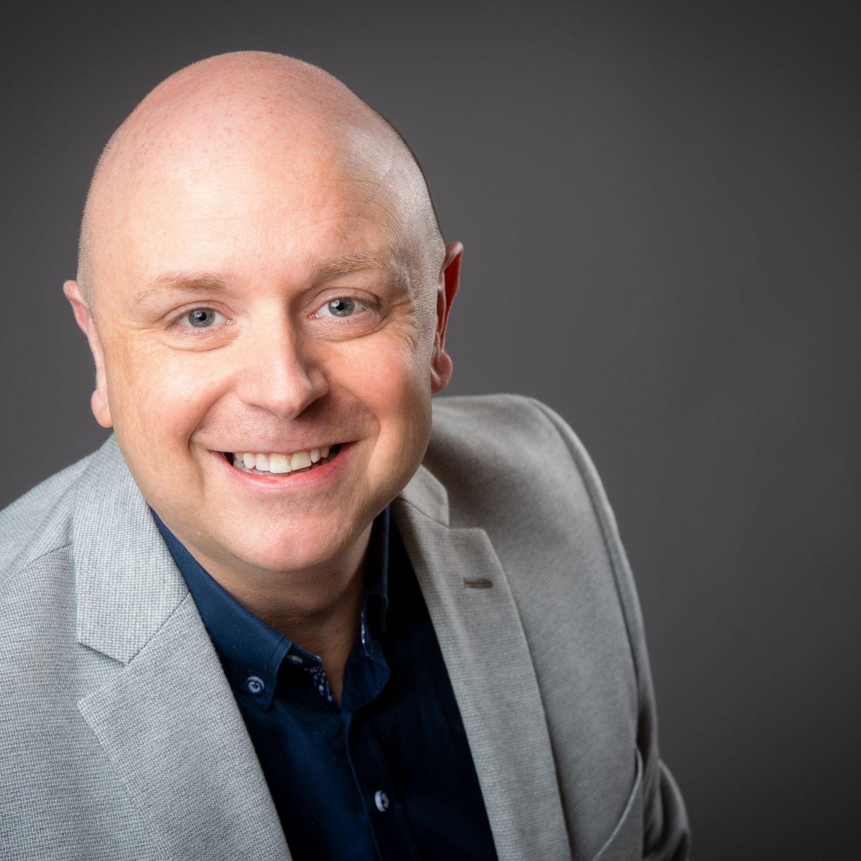 Business headshot of bald man wearing grey suit jacket and orange lanyard