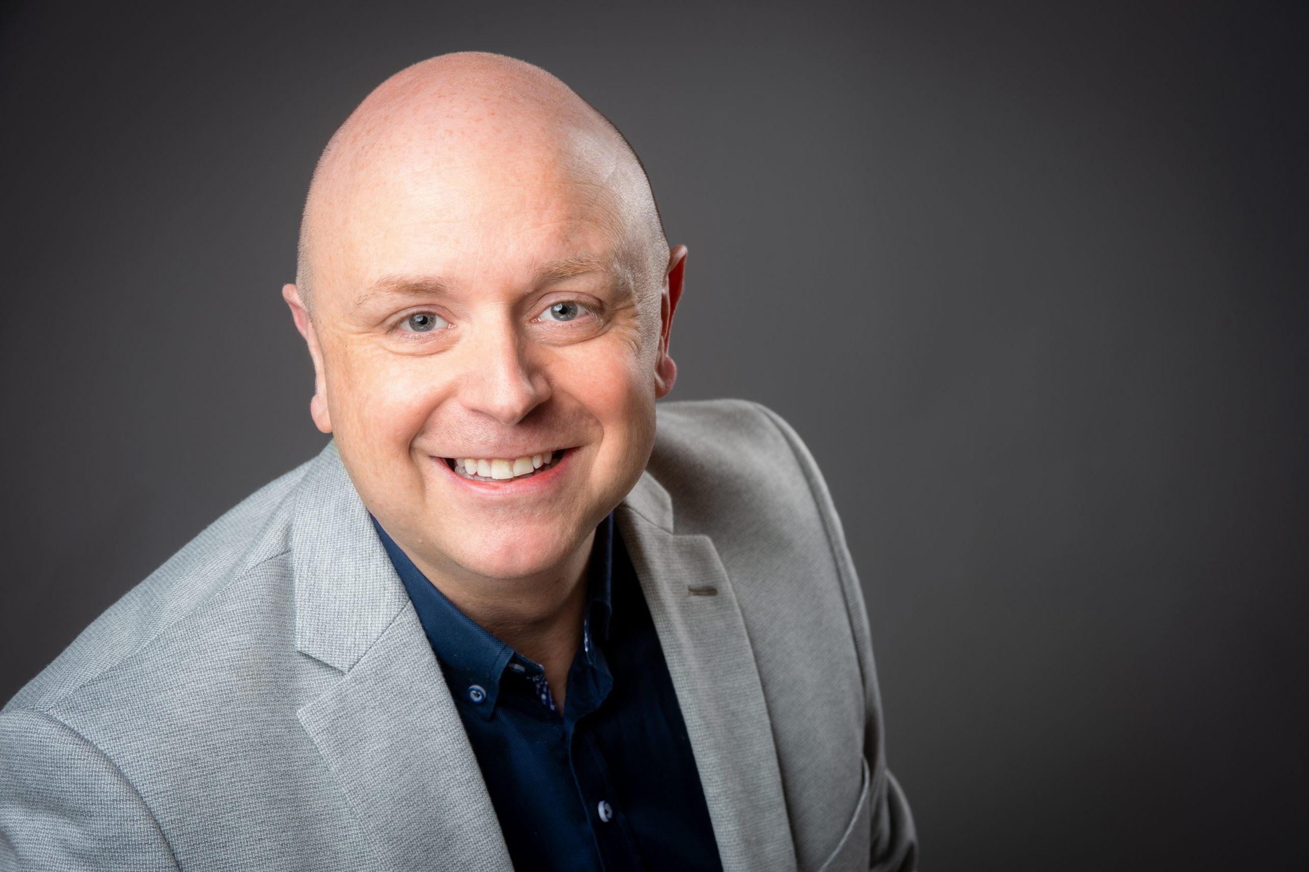 Business headshot of bald man wearing grey suit jacket and orange lanyard